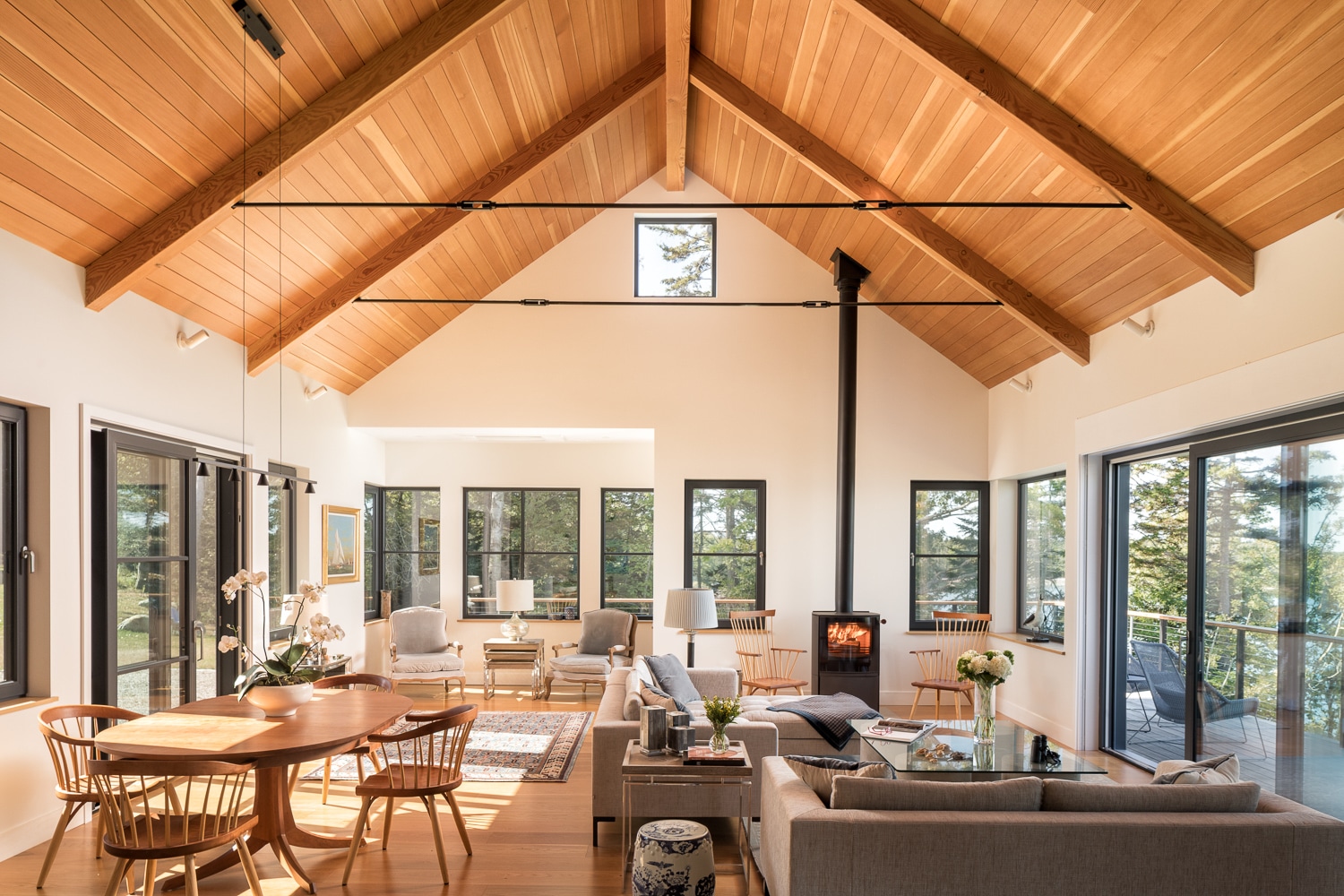 Luxury waterfront great room with vaulted timber ceiling, wood-burning stove, expansive windows, and open-concept living space in a custom Maine home by Warren Construction Group.