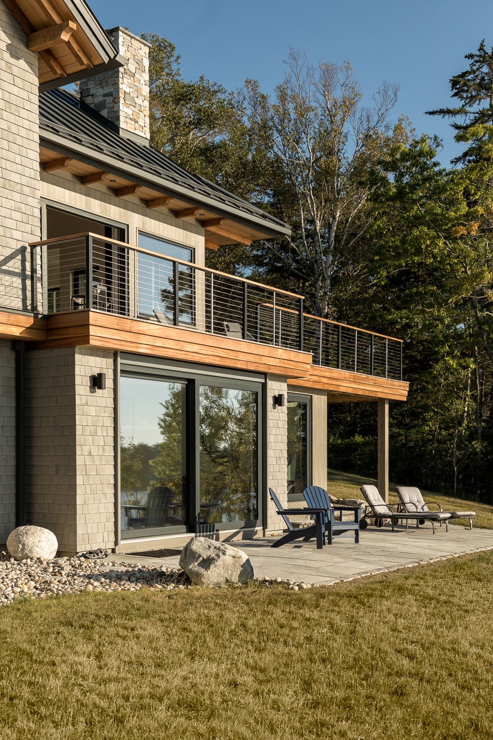 Upper deck with cable rail and lower patio offer views of the ocean from this custom house in Harpswell Maine. Stained cedar shingles and accented wood trim provide a dramatic contrast to the large windows, roof and stone chimney built by Warren Construction Group.