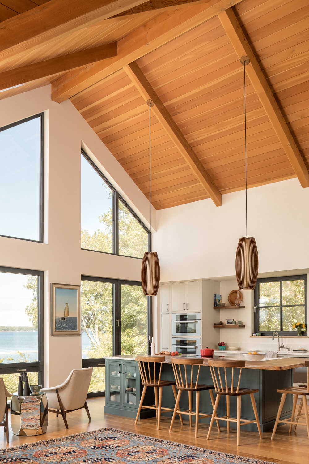 Open-concept luxury kitchen and living area with vaulted timber ceiling, custom cabinetry, large island, and expansive waterfront windows in a custom Maine home by Warren Construction Group.