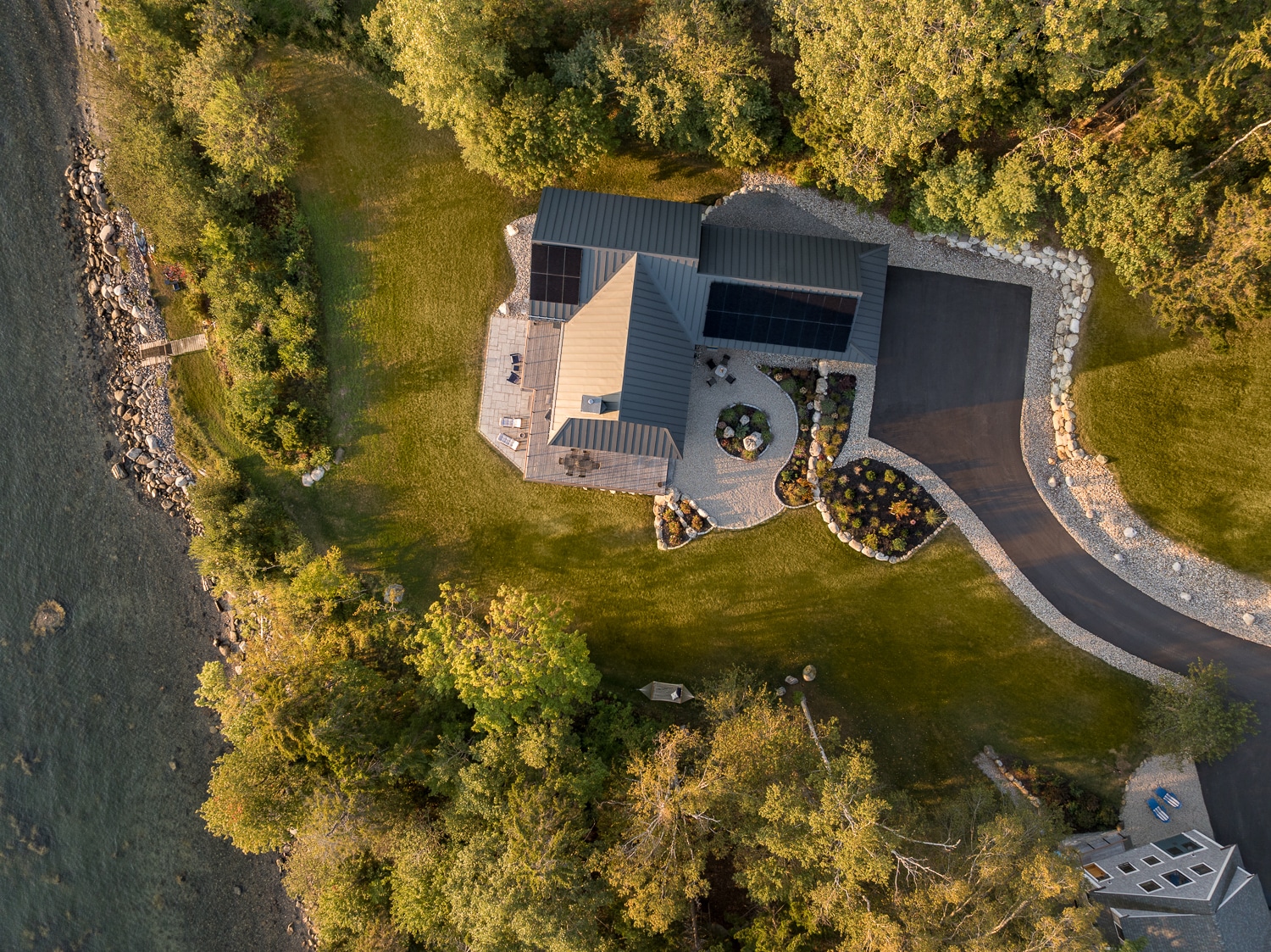 Aerial view of a luxury waterfront home in Maine featuring a standing seam metal roof, rooftop solar panels, landscaped grounds, and private shoreline, built by Warren Construction Group.