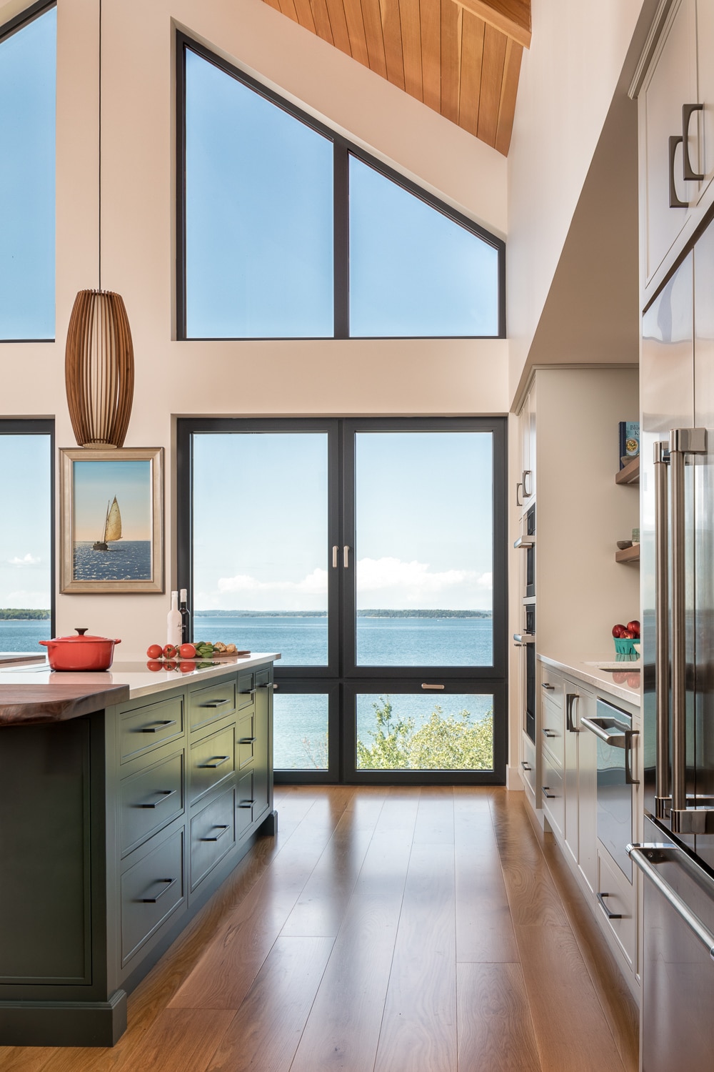 Luxury custom kitchen with floor-to-ceiling windows overlooking the Maine coast, custom cabinetry, vaulted wood ceiling, and large island in a waterfront home by Warren Construction Group.