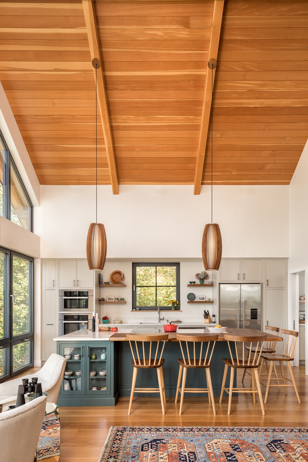 Modern custom kitchen with vaulted wood ceiling, exposed timber beams, custom cabinetry, large island, and floor-to-ceiling windows in a luxury Maine home built by Warren Construction Group.