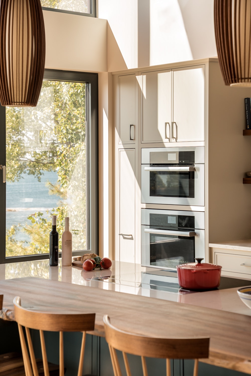 Modern custom kitchen with island, built-in double wall ovens, floor-to-ceiling windows, and waterfront views in a luxury Maine home built by Warren Construction Group.