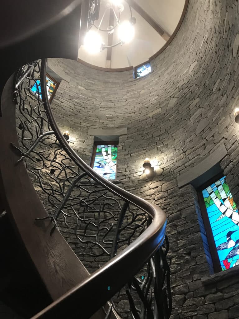 Curved custom staircase with artistic wrought iron railing, natural stone walls, and stained glass windows in a luxury custom home built by Warren Construction Group.