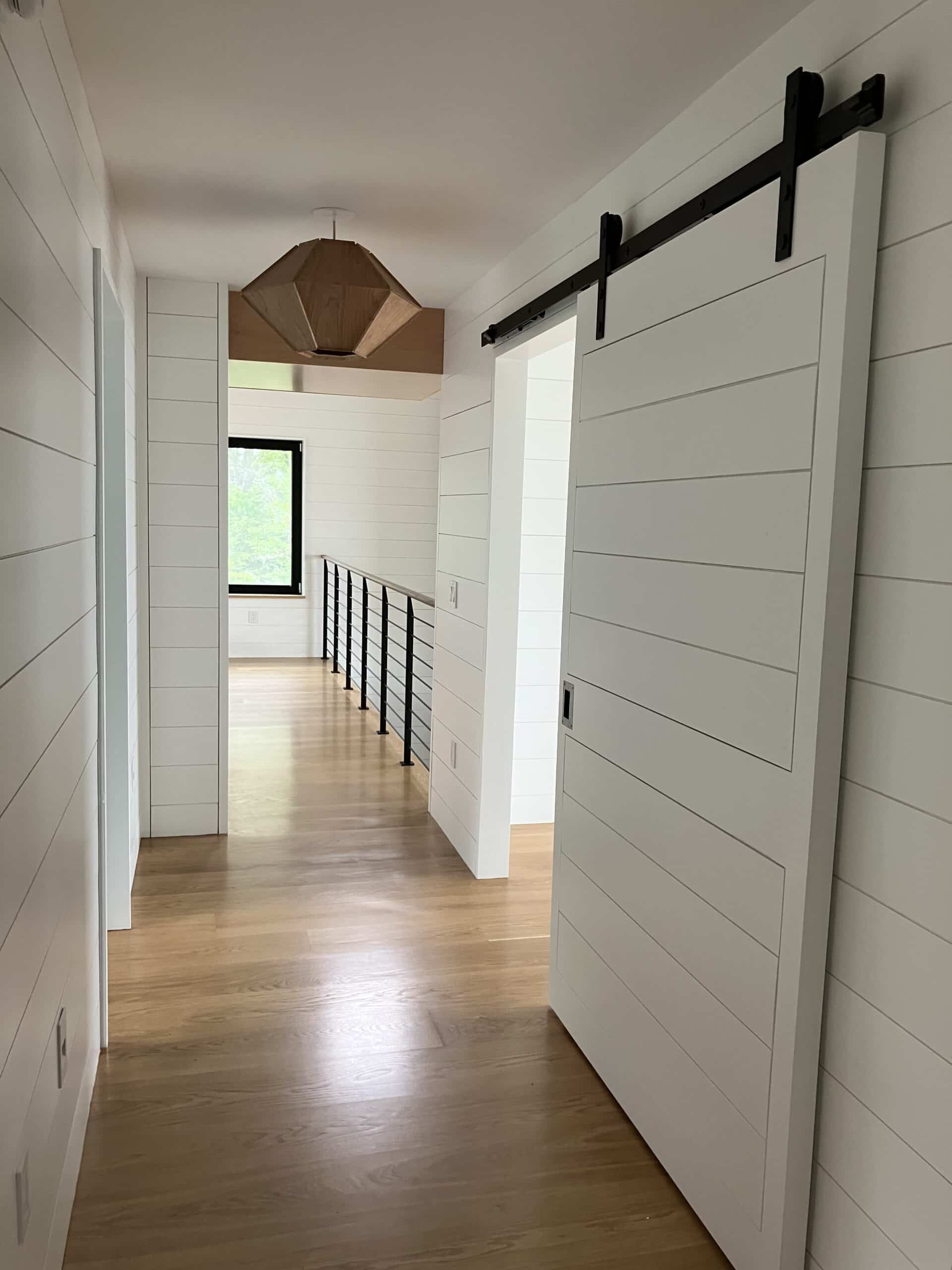 Hallway with a sliding barn door and balcony over looking the first floor. The walls are detailed with white nickel gap boards